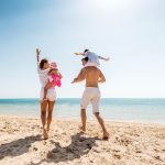 Iloiset vanhemmat nauttivat rannasta lastensa kanssa. Mies kantaa pientä poikaa harteilaan,