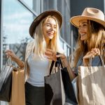 Kaksi hattupäistä nuorta naistaa nauraa yhdessä ostoskasseja kantaen.