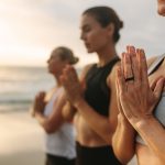 Naiset joogaavat rannalla aikaisin aamulla kädet yhteen liitettyinä ja silmät kiinni