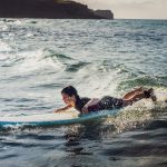 Teini-ikäinen nuori makaa surffilaudan päällä pienessä aallossa