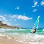 Mies ohjaa purjelautaa rannan suuntaisesti aurinkoisessa säässä.