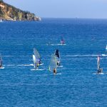 Ryhmä purjelautailijoita etenee merellä aurinkoisessa säässä.