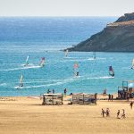 Useita purjelautailijoita rannalta kuvattuna.