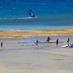 Kaksi purjelautailijaa merellä ja rannalla ihmisiä kulkemassa matalassa vedessä.