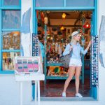 Hattupäinen hymyilevä nainen siniseksi maalatun matkamuistomyymälän ovella katsomassa tuliaisia.