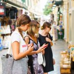 Kolme naista matkapuhelimet kädessä katsomassa kadulla myytäviä matkamuistoja.