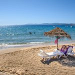 Ihmisiä uimassa kimmeltävässä meressä ja kaksi naista lukemassa aurinkovarjojen alla hiekkarannalla.