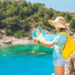 Nainen lierihattu päässään ja keltainen reppu selässään katsoo karttaa meren äärellä.