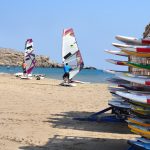 Kaksi miestä säätää purjelautoja aurinkoisella hiekkarannalla.