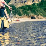 Nainen seisoo rantavedessä uimaräpylät, uimalasit ja snorkkeli käsissään.