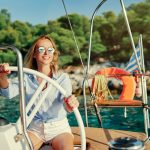 Aurinkolasipäinen nainen pitää kiinni veneen ohjauspyörästä ja hymyilee.