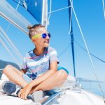 Nuori poika istuu veneen kannella jalat ristissä ja hymyilee iloisesti aurinkolasit päässään.