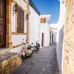 Skootteri ja mopo parkkeerattuna kapealle kujalle valkoisen rakennuksen viereen.