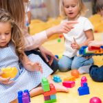 Kolme pientä lasta rakentaa värikkäillä palikoilla aikuisen neuvoessa.