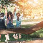 Kolme tyttöä istuu paksun puun oksan päällä kädet ilmassa.