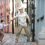 Vanhaan tyyliin pukeutunut hypähtämässä oleva poika pitää isäänsä kädestä kapealla kujalla Riiassa.