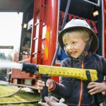 Pieni poika kypärä päässä ruiskuttaa vettä paloauton letkusta.