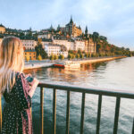 Nainen pitää puhelinta kädessään ja katsoo Tukholman kaupunkinäkymää sillalla.