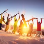 Ryhmä iloisia laskettelijoita nostaneet lasketteluvälineet ilmaan auringon laskiessa.