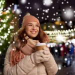 Lämpimästi pukeutunut nuori nainen hymyilee kuppi kädessä jouluvalaistulla kadulla.