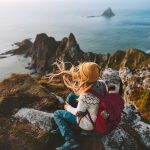 Nainen istuu reppu selässä kallioilla ja ihailee maisemaa merelle.