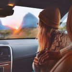 Nainen istuu autossa ja ihailee auringon laskua vuorten taakse.