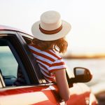 Hattupäinen nainen kurkistaa ulos auton ikkunasta ja katsoo merimaisemaa.