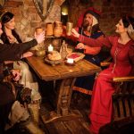 Keskiaikaisiin asuihin pukeutunut seurue kohottaa maljoja puisen ruokapöydän äärellä.