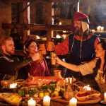 Keskiaikaisiin asuihin pukeutunut seurue kohottaa maljoja illallisella.