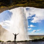 Henkilö seisoo Seljalandsfossin vesiputouksen takana ja levittää kädet ilmaan.