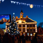 Henkilö ottaa puhelimella kuvaa joulukuusesta sekä jouluvaloin koristellusta kaupungintalosta.