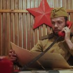 Neuvostoliiton sotilasasuun pukeutunut mies pitää punaista puhelinta korvallaan ja katsoo kansiota.