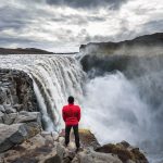 Punapaitainen vaeltaja seisoo Dettifossin vesiputouksen reunalla ja katsoo kuohuja.