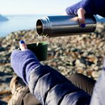 Nainen istuu korkealla kivisessä maastossa vaellusvarusteissa ja kaataa termospullosta kuumaa juotavaa.