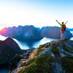 Vuoren päälle kiivennyt nainen nostaa kädet ilmaan keskiyön auringon paistaessa.