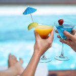 Pariskunta kohottaa värikkäitä drinkkejä altaan reunalla.