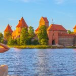 Hattupäinen tyttö istuu rannalla Trakain linnan edustalla ilta-auringossa.