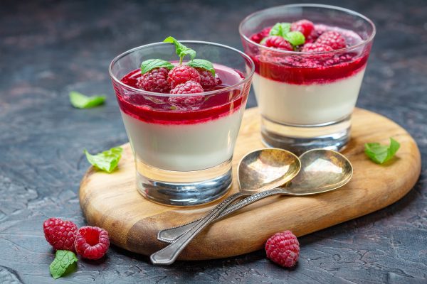Vadelmilla ja hyytelöllä kuorrutetut pannacotta-annokset puisella alustalla, vieressä kaksi lusikkaa.