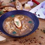 Sipulikeittoa vadissa, päällä leipäpala ja yrttejä. Ympärillä juustoa, sipuleita, mausteita ym. puisella tasolla.