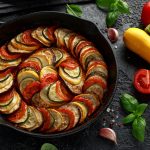 Kasvispannu ylhäältä päin kuvattuna, ympärillä kurpitsoja, tomaatteja, valkosipulin kynsiä ja basilikaa.