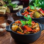 Kaksi pientä pataa täynnä kasvispaistosta puisella tasolla, ympärillä lisukkeita ja mausteita.