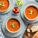 Kolme kulhoa tomaattikeittoa lisukkeineen ylhäältäpäin kuvattuna.