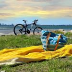 Pyyhe ja laukku rannalla ruohikossa, taustalla pyörä ja kauempana kaupungin siluettia.