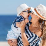 Nainen pitää pientä lasta sylissä, molemmat nauravat, taustalla meri.