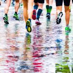 Juoksijoita märällä tiellä, värikkäät asut heijastuvat veden pintaan.