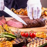 Puolikypsän lihapalan leikkaamista pöydällä, joka on täynnä herkullisen näköisiä kasviksia.