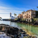 Keltasävyinen kivitalo ja sinivalkoinen majakka kallioisella rannalla.