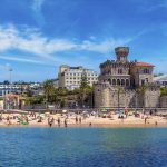 Ihmisiä uimarannalla, taustalla kivimuuri ja vanha rakennus.