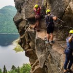 Ryhmä vuorikiipeilijöitä etenee kalliolla, taustalla henkilö köysiradalla korkealla järven yllä.