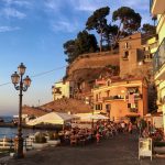 Ihmisiä istumassa ravintoloiden terasseilla Sorrentossa meren rannalla, taustalla erikoisia taloja